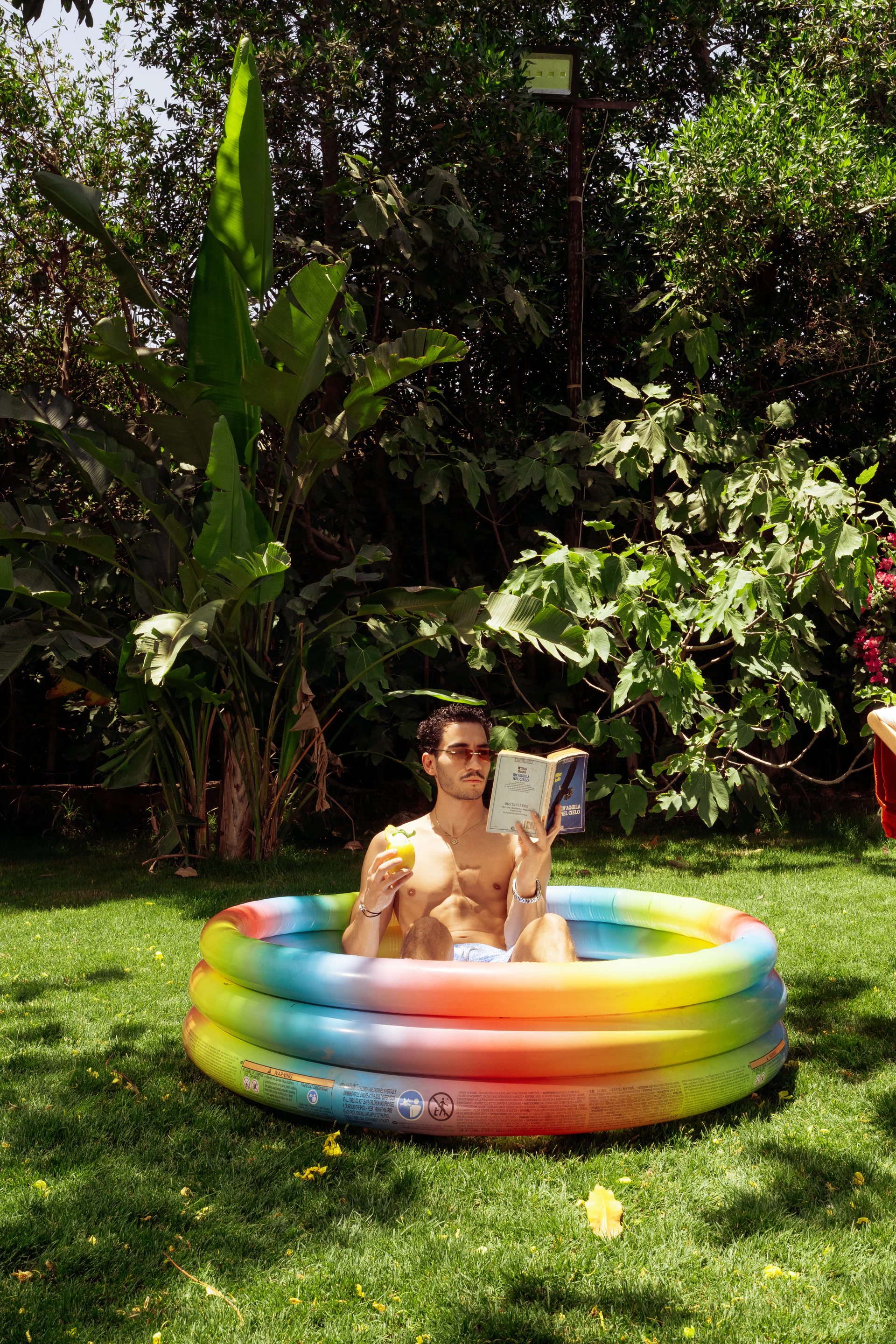 Man reading in a colorful paddling pool while holding a frozen drink