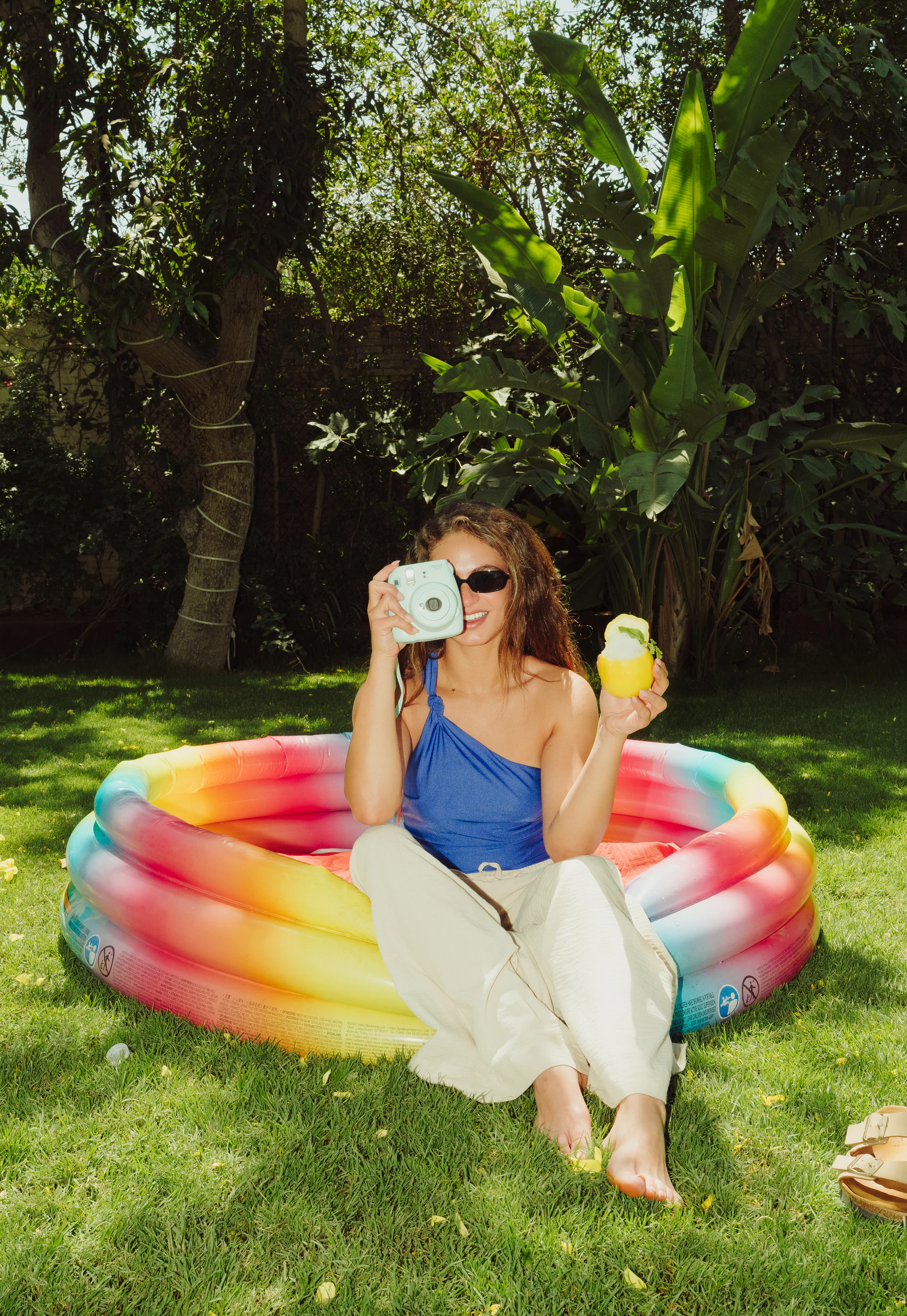 Woman holding a lemon sorbet drink and instant camera beside a colorful pool